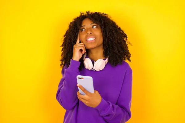 アフリカ系アメリカ人の女性が携帯電話を持ち 黄色の背景で何かを考えている 感情やジェスチャーの概念 — ストック写真