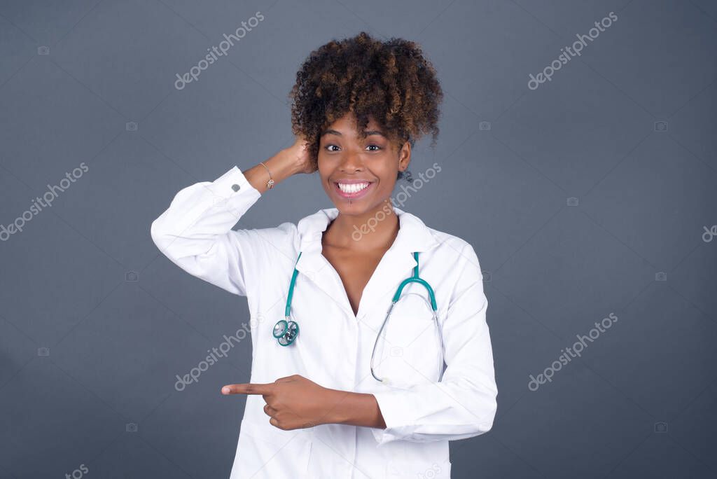 Joyful pretty african american woman doctor  wearing medical uniform demonstrates something. One hand on her head and pointing with other hand.