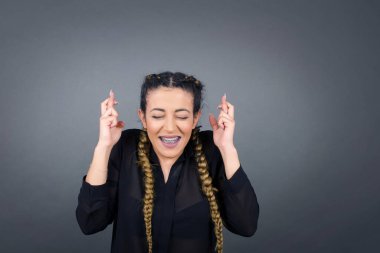 Neşeli genç esmer kadın dişlerini sıkar, parmaklarını çapraz yapar, arzulanan dilekleri yapar, günlük kıyafetler giyer, iyi haberleri bekler, gri duvarın üzerinde durur.