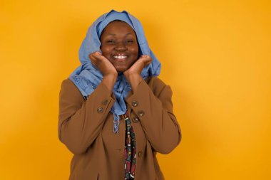 Hayalperest genç Afrikalı Amerikalı Müslüman kadın ellerini çenesinin altında tutuyor, mutlu bir ifadeyle bakıyor, dişlek gülüşü var, sarı arka planda izole edilmiş.