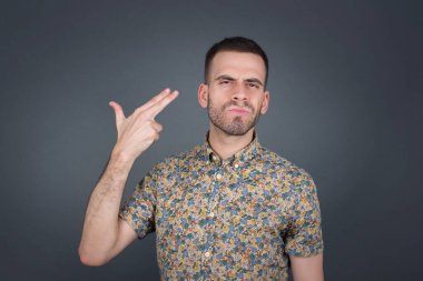 Mutsuz sarışın genç erkek silah atışı taklidi yapıyor intihar jesti yapıyor iki parmağını şakaklarda tutuyor günlük kıyafetler giymiş şaşkın yüz ifadesi içeride duruyor..