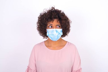 Yüz maskesi takan ve sıradan giyinen bir kadın beyaz stüdyo arka planında izole edilmiş bir şekilde yüzünü çeviriyor.