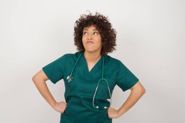 Sağlık üniforması giyen, bir şeyler düşünen güzel Arap doktor kadının belden yukarı pozu. Ellerini kalçaya koy, yüz ifadenden şüphe et. Gri duvara yaslanmış bir şey olup olmadığını merak ediyorum..