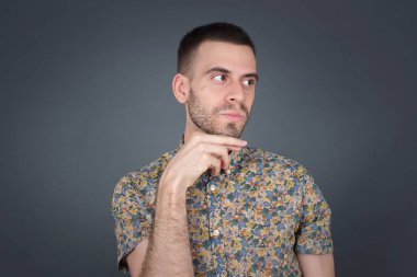 Hayalperest bir erkek, hoş bir yüz ifadesi var, günlük kıyafetler giyiyor, yan bakıyor, elini çenesinin altında tutuyor, hoş bir şeyler düşünüyor, gri arka planda poz veriyor..