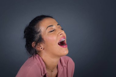 Neşeli bir kadın iyi bir şakaya gülüyor ve eğleniyor ya da günlük kıyafetler giyiyor, gri duvara yaslanıyor..