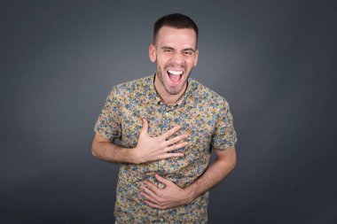 Neşeli Avrupalı adam ellerini çapraz tutuyor, güzel şakalara gülüyor, günlük kıyafetler giyiyor ve gözlük takıyor, içeride duruyor. Uzun saçlı mutlu genç adam gri duvara poz veriyor..