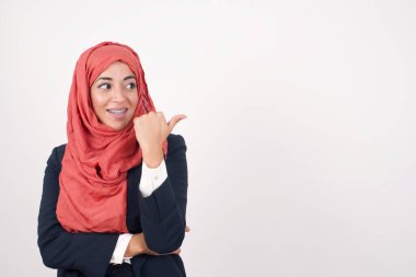 Omg konsepti. Şaşırmış güzel Müslüman kadın siyah ceket giyer ve şaşkın bir ifadeyle tesettürlü, geniş bir şekilde gözlerini ve ağzını açar, baş parmağıyla işaret eder, gri arka planda garip bir şey gösterir. Reklam kavramı.