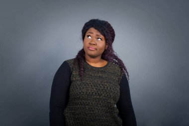 Kafası karışık Afro-Amerikan kadını, sıradan kıyafetler içinde, kaşlarını çatar, dudaklarını ısırır, kaşlarını kaldırır, yukarı bakar, sorunları olur, şık kıyafetler giyer, gri arka planda soyutlanır, pişman görünür..