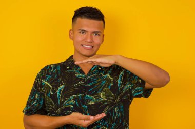 Genç, yakışıklı, İspanyol bir adam izole edilmiş arka plan üzerinde gündelik giysiler giyiyor. Ellerinde büyük ve büyük boy işareti, ölçü sembolü var. Kameraya bakıp gülümsüyor. Ölçüm konsepti.