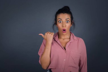 Genç ve neşeli bir kadının başparmak ucunda yatay görüntüsü gri arka planda izole edilmiş boş bir duvara işaret ediyor..