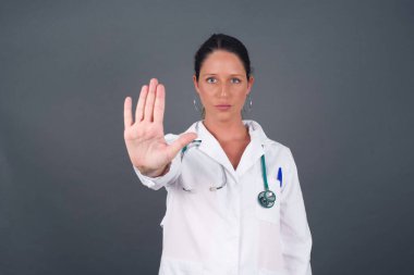 Gri arka planda izole edilmiş genç, beyaz, güzel doktor kadın avuç içiyle dur hareketi yapıyor. Gri arkaplan üzerinde izole edilmiş yüz üzerinde negatif ve ciddi jest içeren uyarı ifadesi.
