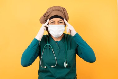 Yeşil üniformalı genç, Müslüman doktor kadın ciddi bir bakışla bir fikre odaklanıyor, her iki işaret parmağı alnını göstererek ve kameraya bakarak düşünüyor..