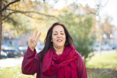 Olgun Arap kadın parkta duruyor ve parmaklarını dört numaraya doğrultuyor. Kendinden emin ve mutlu bir şekilde gülümsüyor..