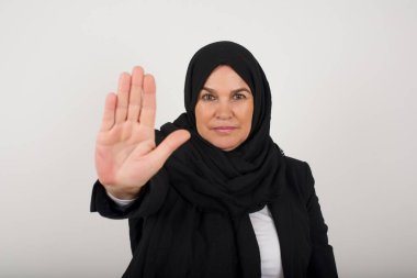 Siyah tesettürlü genç müslüman kadın avuç içiyle el hareketini durduruyor. Gri arkaplan üzerinde izole edilmiş yüz üzerinde negatif ve ciddi jest içeren uyarı ifadesi.