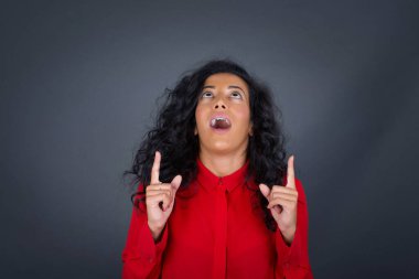 Kaygısız esmer, kıvırcık saçlı, kırmızı tişörtlü, her iki parmağı işaret eden, gündelik kıyafetler giymiş, geniş bir tebessüme sahip, gri arka planda soyutlanmış, kaygısız bir kadın. Şuraya bakın, lütfen..