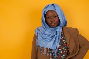 Afro-Amerikalı neşeli Müslüman kadının stüdyo çekimi elini kalçasında tutuyor, geniş bir gülümsemeyle, arkadaşlarının doğum gününde hazırladığı sürprizi bekliyor, sarı arka planda izole edilmiş..