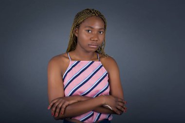 Endişeli ve korkmuş genç Afrikalı Amerikalı kadın gri arka planda dururken kollarını kavuşturuyor.