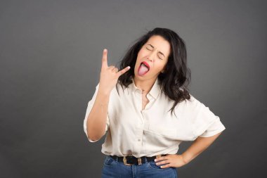 Güzel esmer kadın, boynuzlu jestlerle sıradan giyinmiş. Gri arkaplanda duruyor. 