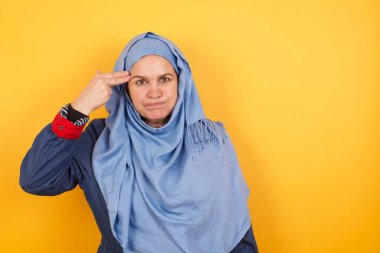 Mutsuz, orta yaşlı, tesettürlü Müslüman kadın silah atışı taklidi yapıyor intihar jestini iki parmağında tutuyor.