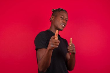 Hey sen, bang. Neşeli ve karizmatik yakışıklı, siyah tişörtlü, kırmızı arka planda siyah tişörtlü Afro-Amerikan bir adam, kameraya mutlu bir şekilde ve arsız bir şekilde beyaz duvarın üzerinde poz veriyor.