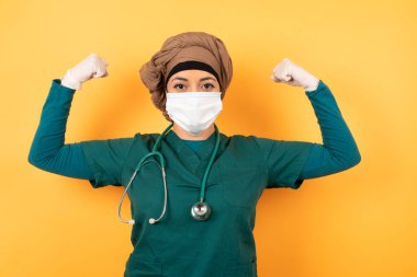 Yüz maskeli kadın doktorun belden yukarı pozu kollarını kaldırarak zaferden emin olduğunu, güçlü ve bağımsız göründüğünü, kameraya olumlu gülümsediğini, arka planda durduğunu gösterir..