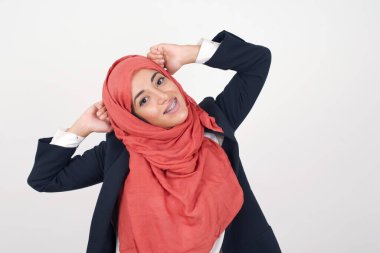 Güven ve cinsel ilişki konsepti. Güzel Müslüman bir kadının portresi siyah ceket ve tesettür giyer. Kendine güvenen bir ifadeyle gülümseyerek ellerini başının üstüne koyar. Gri arkaplanda duruyor.