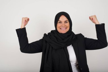 Siyah tesettürlü müslüman bir kadının belden yukarı vuruşu kollarını kaldırarak zaferden emin olduğunu, güçlü ve bağımsız göründüğünü, kameraya olumlu gülümsediğini, gri arka planda durduğunu gösterir. Spor konsepti.