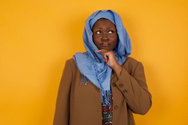 Düşünceli genç Afrikalı Amerikalı Müslüman kadın elini çenesinin altında tutuyor, düşünceli bir şekilde yukarı bakıyor, gündelik kıyafetler giyiyor, sarı arka planda duruyor ve mesajınız için boş yer var.