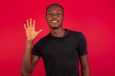 Genç Afro-Amerikan adam duvarın önünde duruyor ve parmaklarını beş numaraya doğrultuyor ve gülümsüyor kendinden emin ve mutlu..