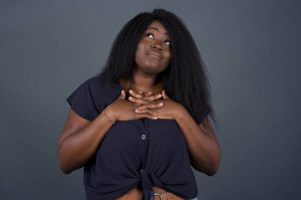Happy African American woman standing and smiling isolated on gray studio background has hands on chest near heart. Young emotional woman. Human emotions, facial expression concept. Front view.