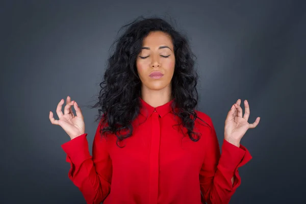 Beautiful young brunette woman with curly hair wearing red shirt doing yoga in the morning outdoors after waking up, keeping eyes closed, holding fingers in mudra gesture.