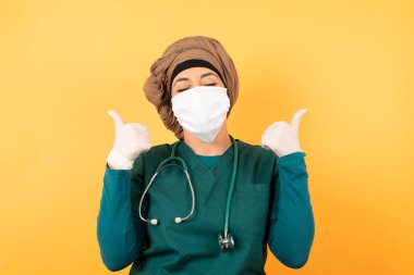 Yüzü maskeli Arap kadın doktorun yakın çekimi arka planda izole edilmiş olumlu içerik ve mutluluk duygularıyla baş parmaklarını gösteriyor. 