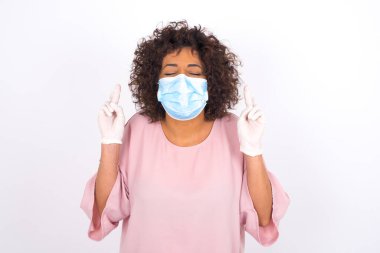 Yüz maskesi takan ve günlük kıyafetler giyen bir kadın beyaz stüdyo arka planında dilek tutuyor.