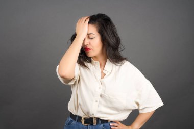 Başı ağrıyan, sıradan giyinmiş güzel esmer kadın. Gri arkaplanda duruyor. 
