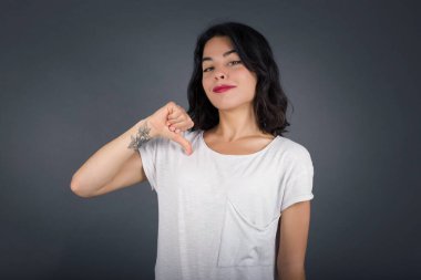 Neşeli, güzel Avrupalı esmer kadının yakın çekimi neşeli, memnun ve kendinden emin görünüyor. Başparmağıyla işaret ediyor. Gri stüdyo arka planında izole edilmiş.. 