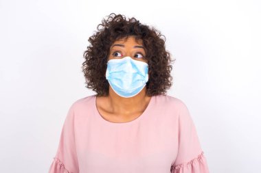 Yüz maskesi takan ve sıradan giyinen bir kadın beyaz stüdyo arka planında izole edilmiş bir şekilde yüzünü çeviriyor.