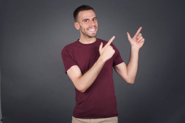 Olumlu ifadeli genç bir adam, sıradan giyinmiş, promosyon metnin ya da reklamın için boş fotokopi alanında ön parmaklarıyla işaret ediyor. Sevimli adam evde tek başına poz veriyor.