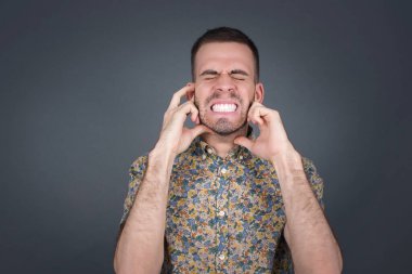 Bu sinir bozucu sesi çıkarmayı kes! Mutsuz genç erkek vesikalığı endişeli yüz ifadeleri takınıyor, parmaklarıyla kulaklarını tıkıyor, üst katındaki komşulardan gelen gürültüden rahatsız oluyor..