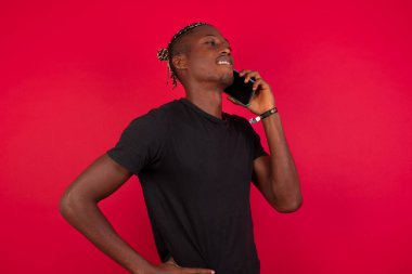Yaşam Tarzı Konsepti - Kırmızı arka planda siyah tişört giyen başarılı, neşeli Afro-Amerikan bir adamın portresi arkadaşıyla cep telefonunda konuşuyor. Kırmızı pastel stüdyo arka planı. Boşluğu Kopyala.