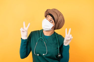 Yüz maskesi takmış neşeli Arap kadın doktor iki eliyle barış ya da zafer işareti yapar, rahat kıyafetler giyer, serin hisseder dişli bir gülümsemesi vardır, arka planda izole olur.. 