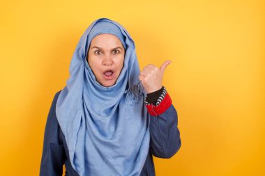 Başparmağıyla tesettürlü orta yaşlı Müslüman bir kadının yatay çekimi arka planda izole edilmiş boş bir duvara işaret ediyor.. 