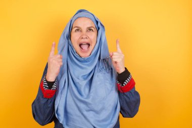 Hoş, orta yaşlı, tesettürlü, olumlu ifadeli, her iki işaret parmağıyla işaret eden, gündelik kıyafetler giymiş, geniş bir tebessüme sahip, geçmişinde izole edilmiş Müslüman bir kadın. Şuraya bakın, lütfen..