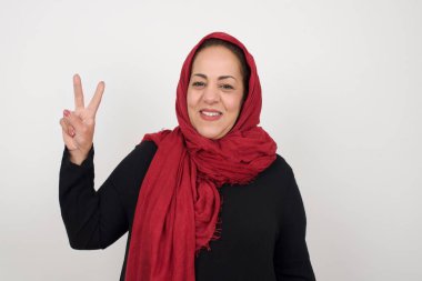 Olgun Müslüman bir kadın gri duvara yaslanmış iki numaralı parmaklarını gösterirken kendinden emin ve mutlu bir şekilde gülümsüyor. Stüdyoda poz veren başörtüsü takan orta yaşlı bir Arap kadın.