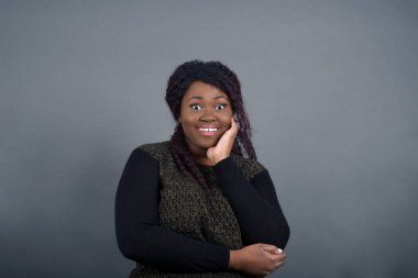 Şaşırtıcı derecede güzel Afro-Amerikan kadını, günlük giysiler içinde, ağzı açık, ağzı açık, ağzı açık, eli yüzün yanında şaşırmış görünüyor. Olumlu duygu yüz ifadesi vücut dili.