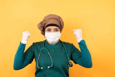 Güçlü Arap kadın doktor portresi. Yüz maskesi takmış, yumruklarını sıkmış, neşe ve heyecanla haykırıyor. Zafer, başarı ve başarı kavramı.