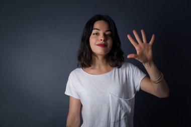 Gri duvara yaslanmış genç bir kadın, mutlu ve kendinden emin gülümserken beş numarayı gösterip işaret ediyor..