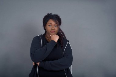 Düşünceli gülümseyen Afrikalı Amerikalı kadın portresi elini çenesinin altında tutuyor, doğrudan kameraya bakıyor, ilgi çekici bir şey dinliyor, sıradan giyiniyor, gri duvara karşı poz veriyor.. 