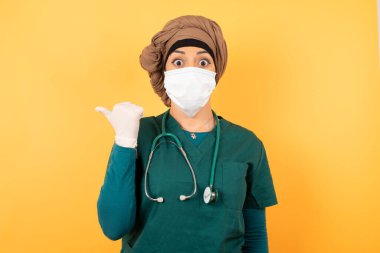 Yeşil üniformalı güzel Müslüman bir kadının yatay görüntüsü, başparmağıyla işaret ediyor, arka planda izole edilmiş boş bir duvara işaret ediyor. Şuna bir bak..