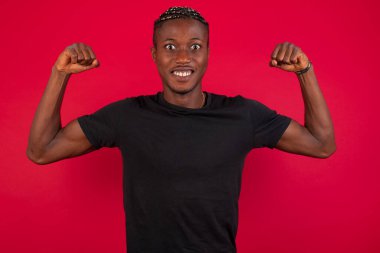 Waist up shot of African American man wearing black t-shirt over red background raises arms to show his muscles feels confident in victory, looks strong and independent, smiles positively at camera, stands against red background. Sport concept. clipart