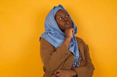 Düşünceli gülümseyen Afrikalı Amerikalı Müslüman kadının portresi elini çenesinin altında tutuyor, yan bakıyor, ilgi çekici bir şey düşünüyor ya da merak ediyor sarı stüdyo arka planında gelişigüzel poz veriyor. Karar alma kavramı.